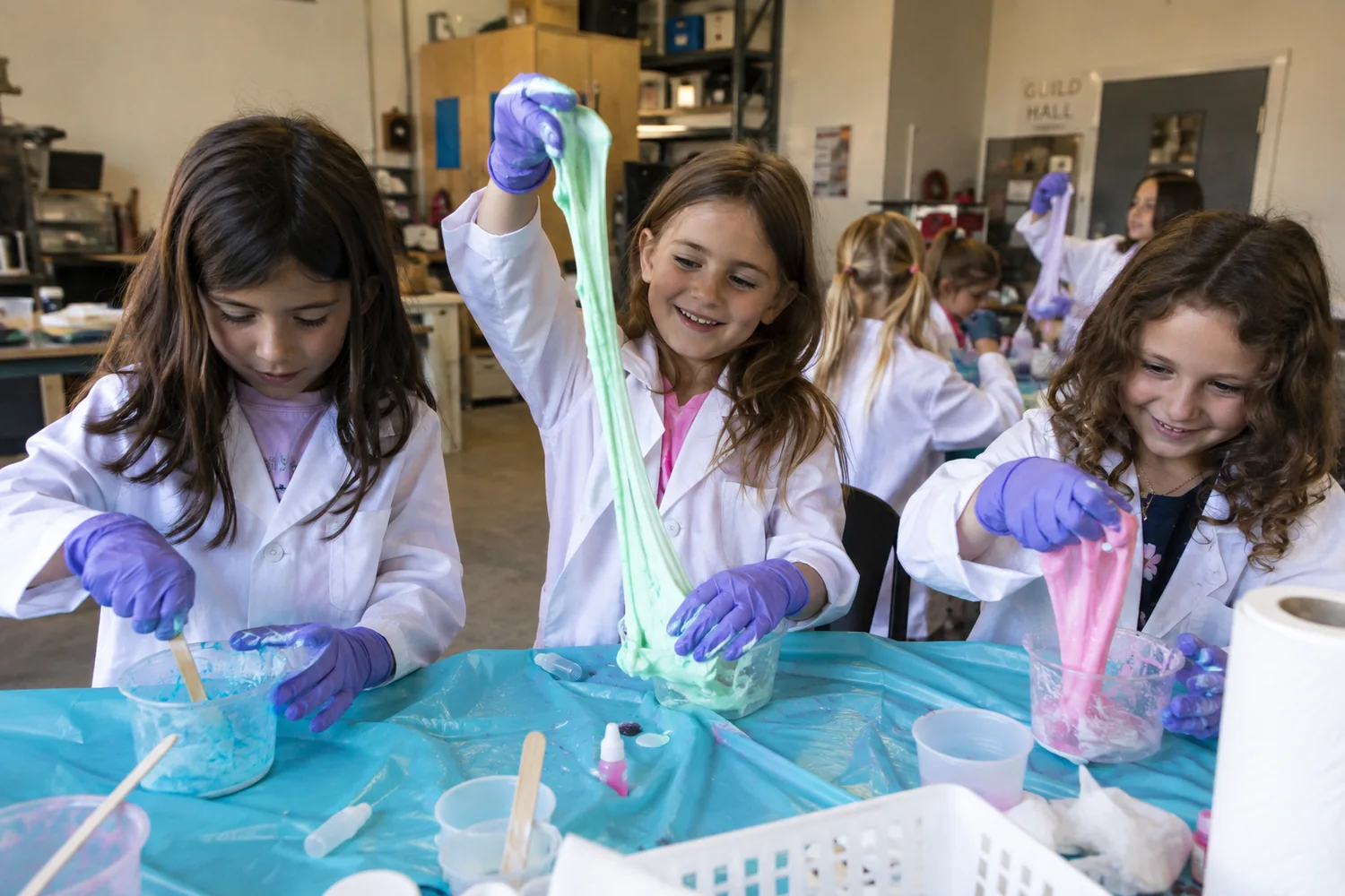 Three young learners in lab coats stretching colorful slime over a covered table