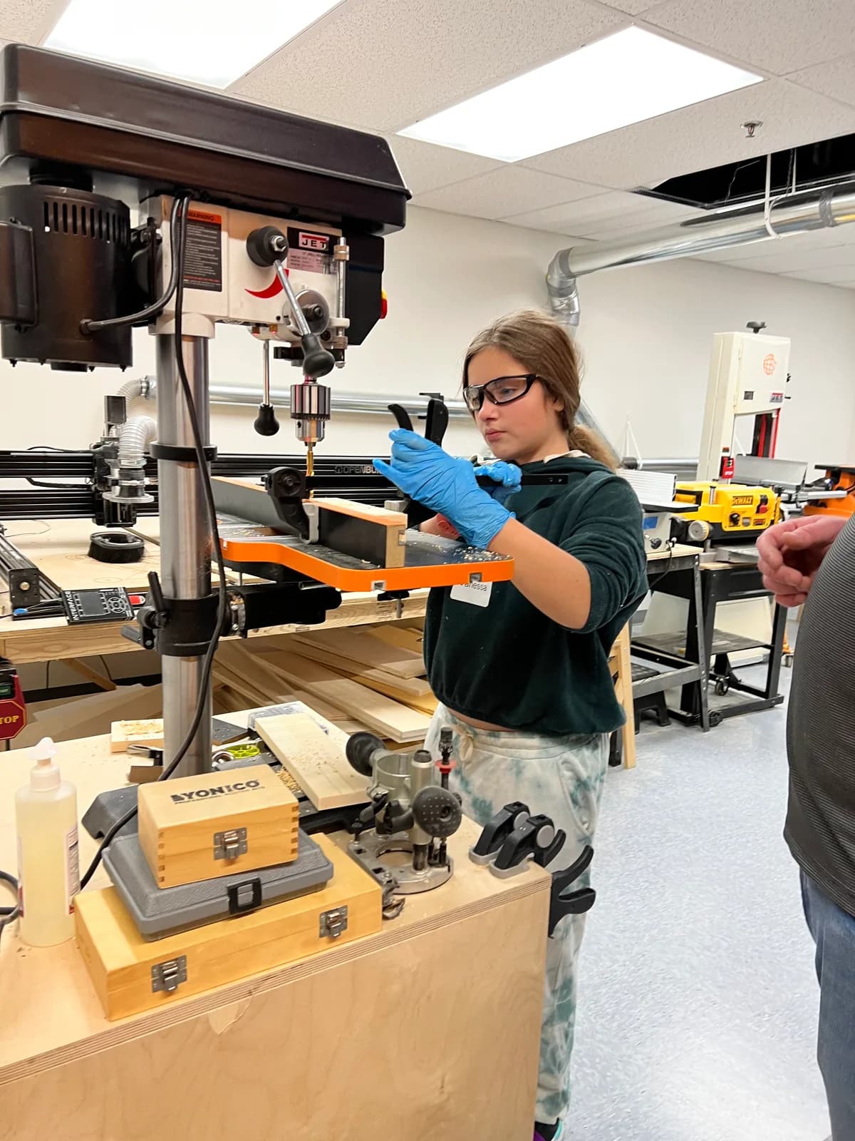 A learner wearing safety glasses positioning a clamped workpiece at a drill press