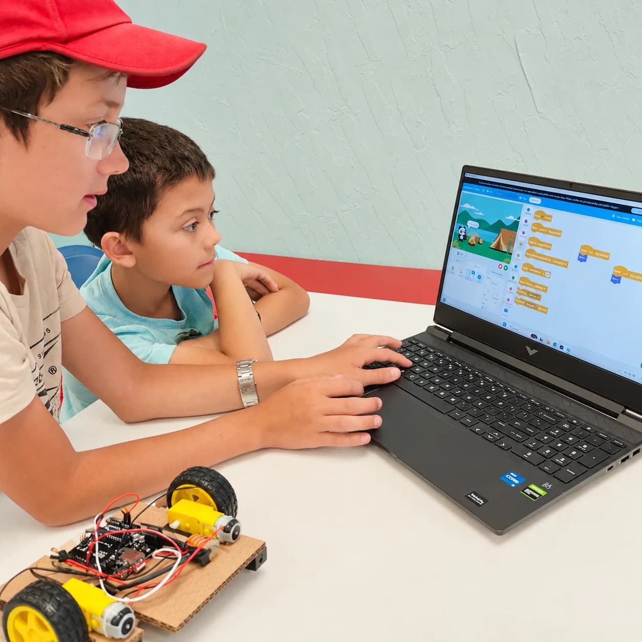 A young learner watching a mentor work with visual coding beside a robot